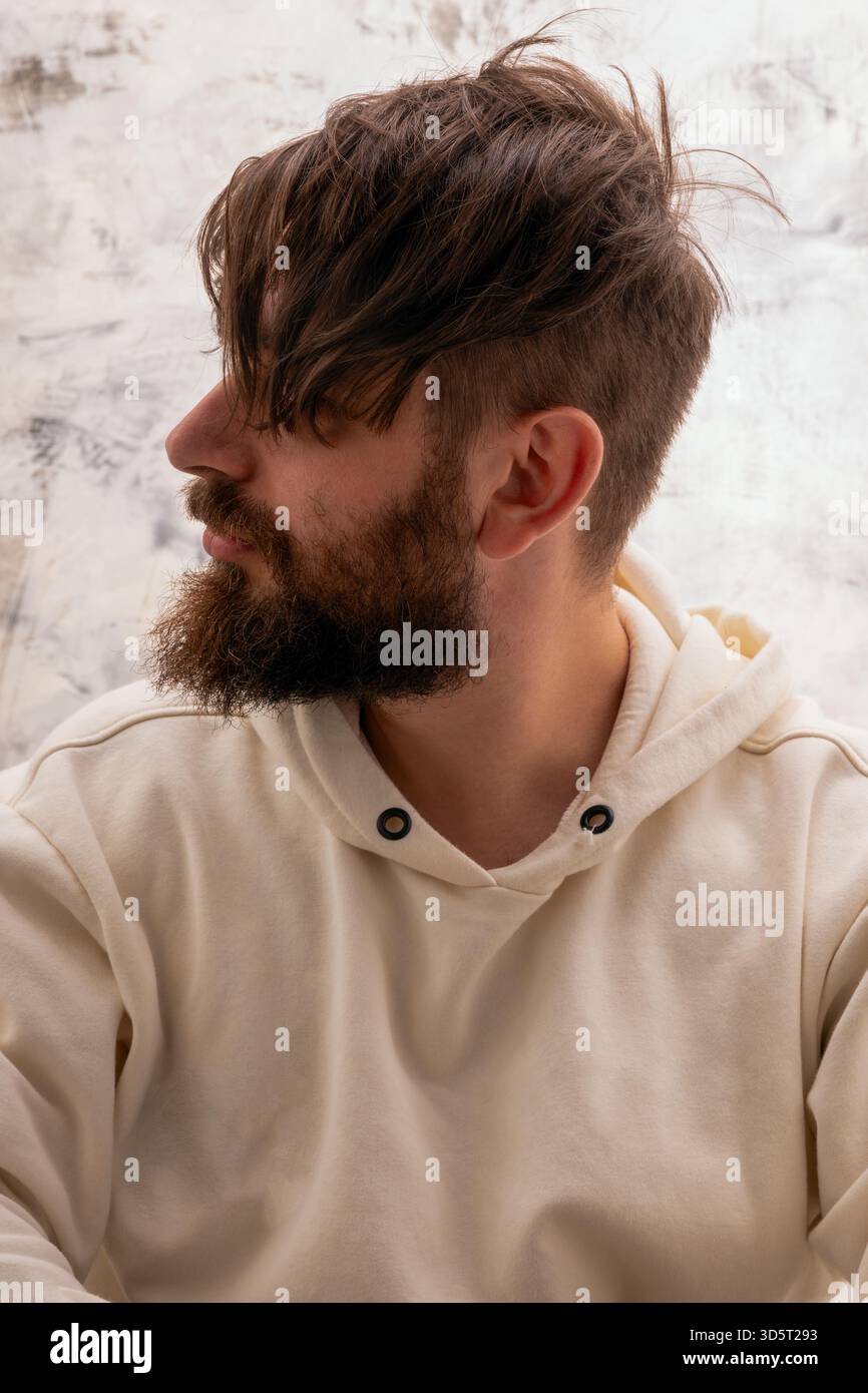 Vista del profilo di un giovane uomo con barba e capelli ruvidi, che indossa una felpa con cappuccio bianchissima su uno sfondo neutro e testurizzato. Foto Stock