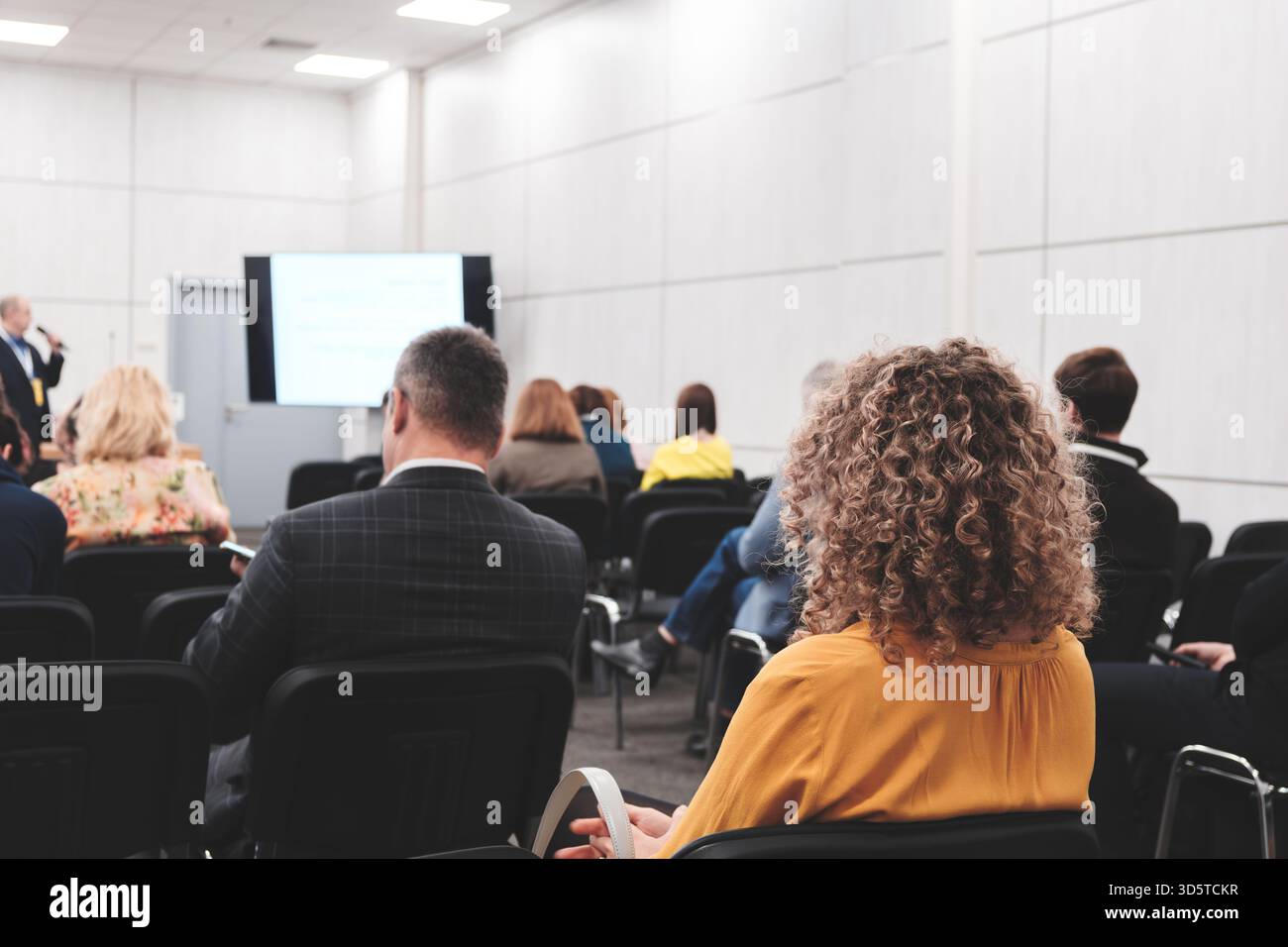 Una donna d'affari siede in una sala conferenze piena di persone. Un altoparlante maschio in una tuta si trova di fronte a uno schermo di proiezione con un microfono. Affari Foto Stock