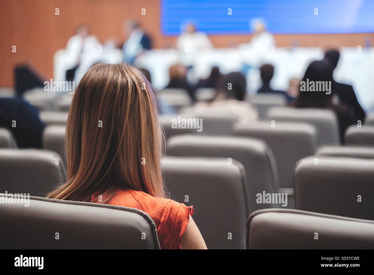 Una donna d'affari siede in una sala conferenze piena di persone. Un altoparlante maschio in una tuta si trova di fronte a uno schermo di proiezione con un microfono. Affari Foto Stock