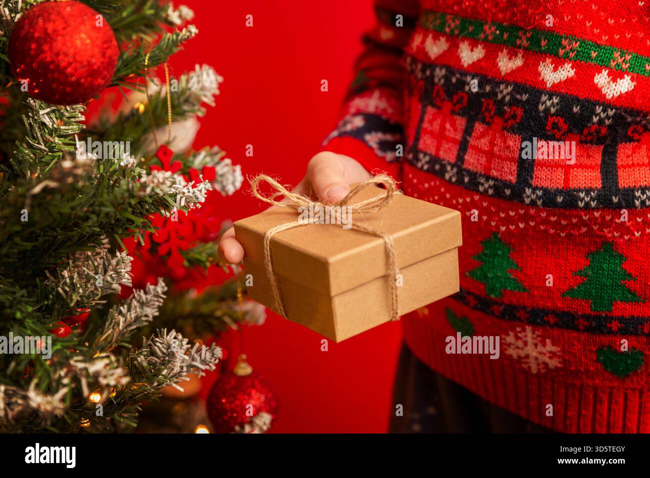 Bambino che tiene un regalo avvolto accanto a un albero di Natale con ornamenti rossi, bacche e rami invernali festivi Foto Stock