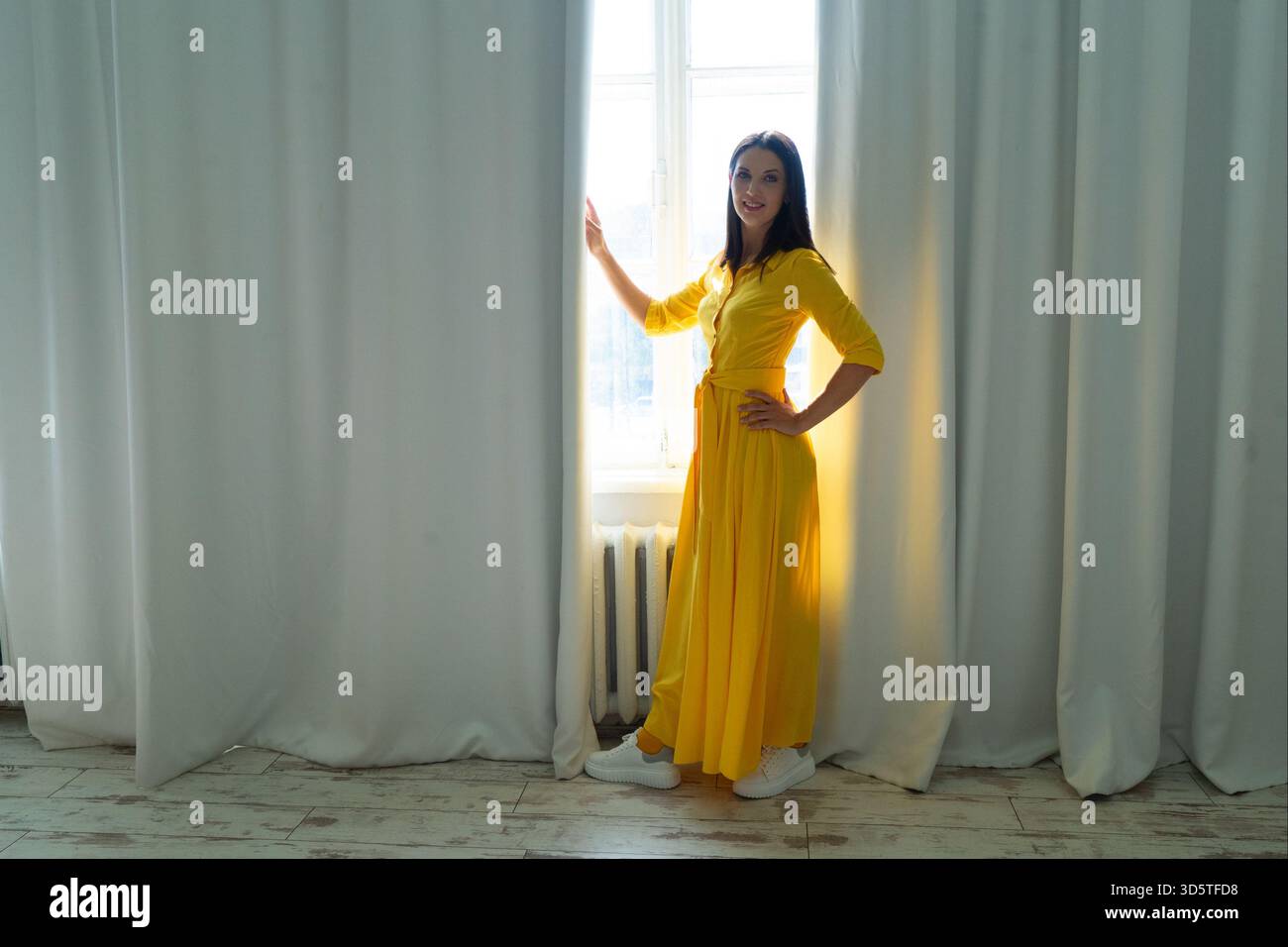 Una donna allegra vestita con un vivace abito giallo posa graziosamente e felicemente vicino alla finestra luminosa Foto Stock