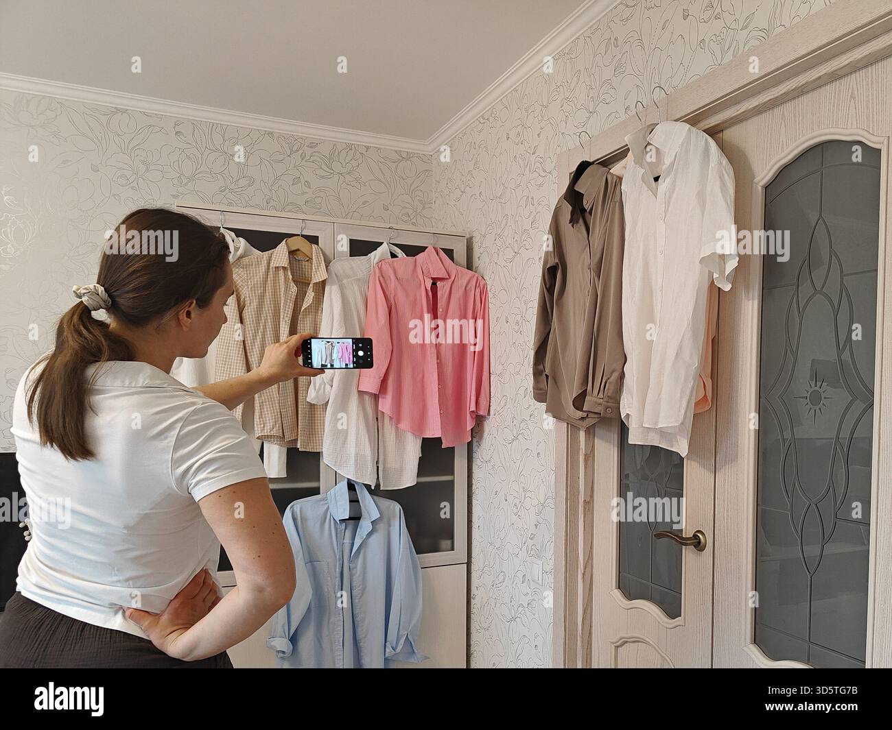 Donna che fotografa capi colorati esposti su un rack in una stanza ben illuminata, mettendo in evidenza le scelte di moda e gli elementi di design degli interni Foto Stock