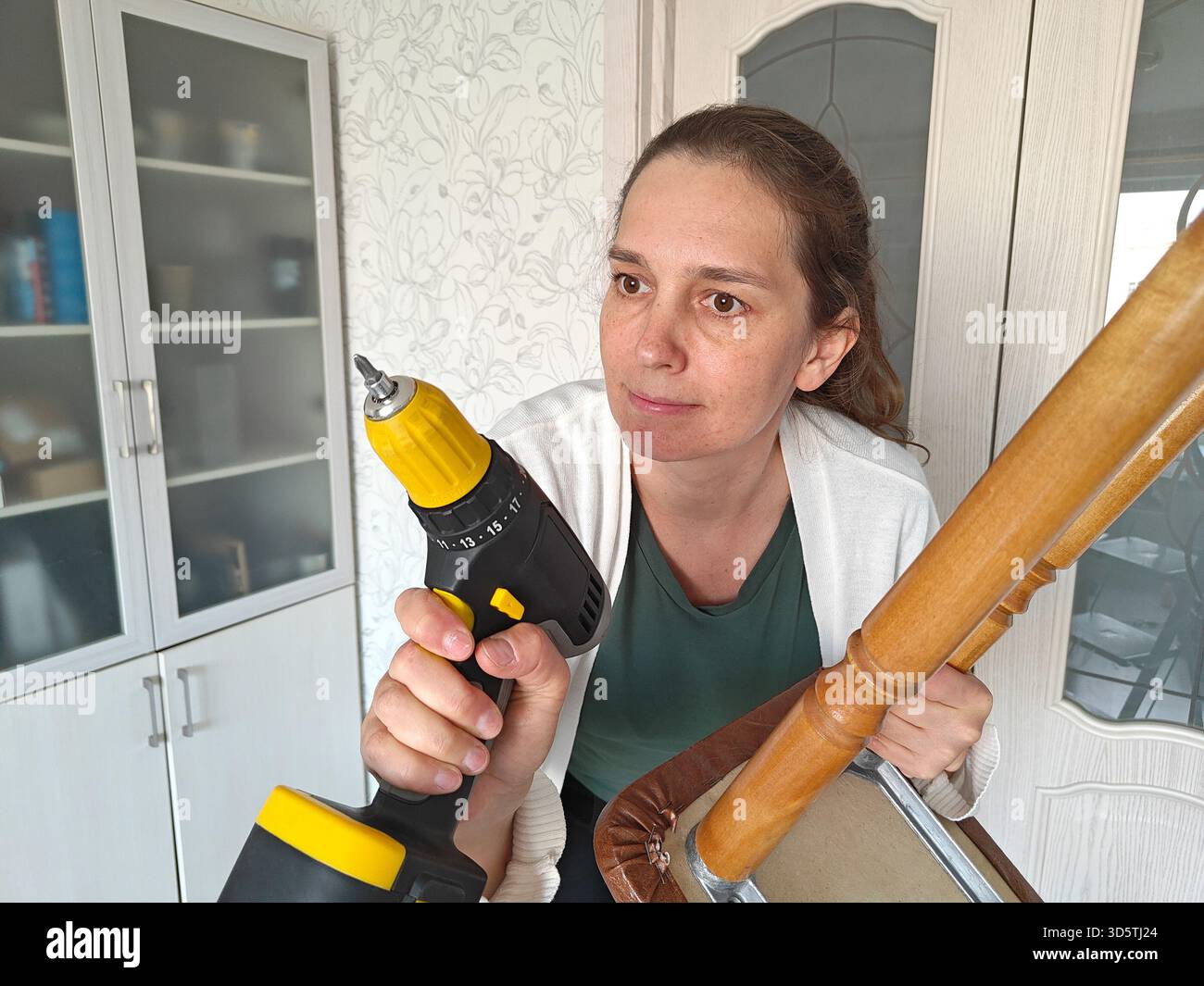 Una donna appassionata di fai da te sta utilizzando un trapano elettrico per riparare una sedia in legno in una stanza ben illuminata, dimostrando le sue abilità e dedizione al miglioramento della casa Foto Stock