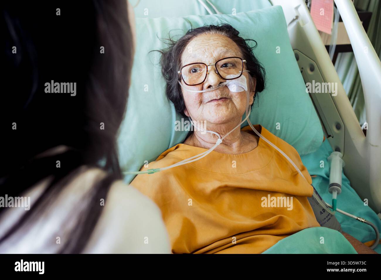Paziente anziana malata del sud-est asiatico in letto d'ospedale con supporto di ossigeno che indossa occhiali sembra felice e appassionata di essere visitata dalla famiglia Foto Stock