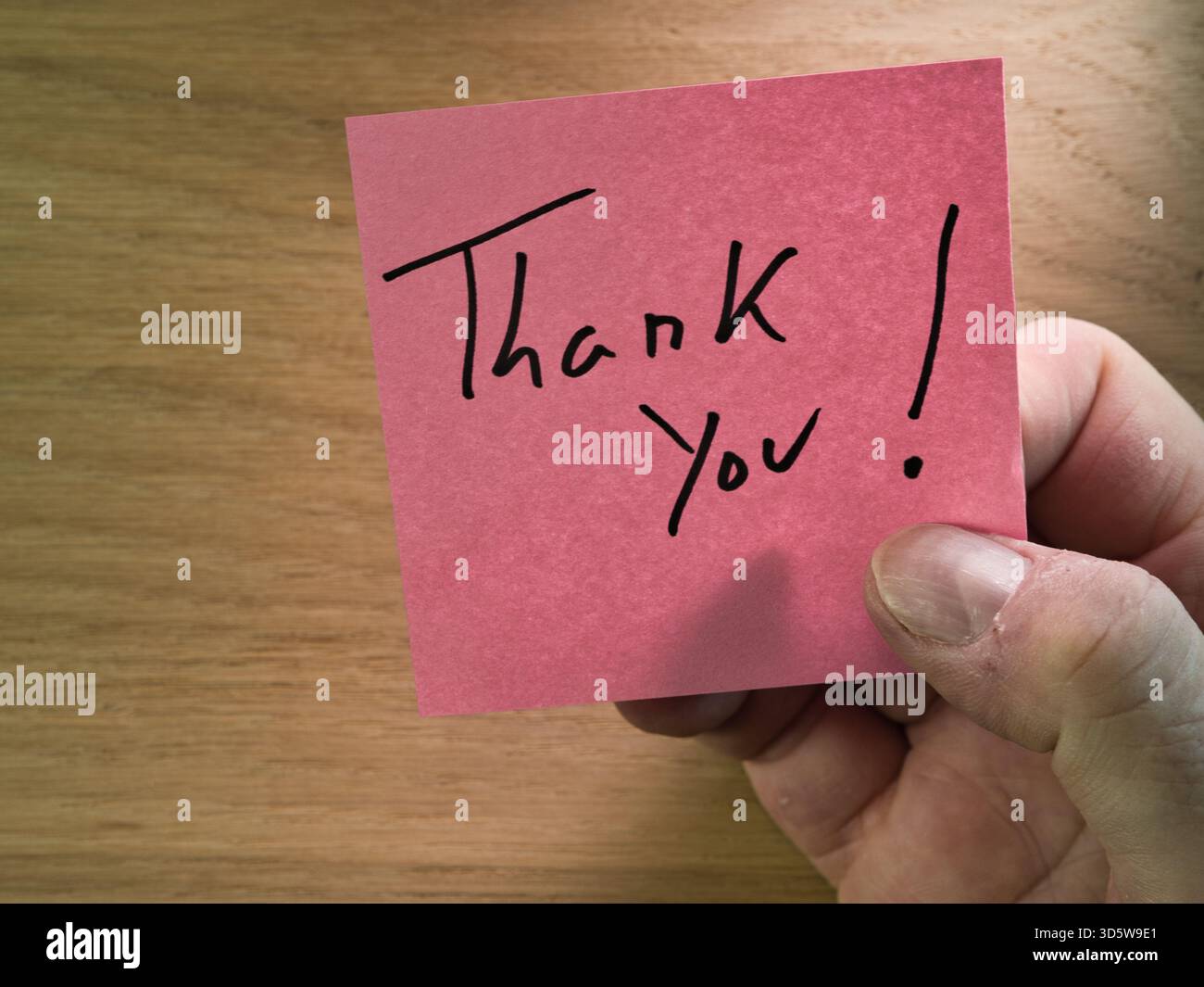 Nota di ringraziamento tenuta a mano su legno - mano che tiene una nota adesiva rosa vivace con "Grazie!" scritto a mano, su un caldo sfondo di legno. A c Foto Stock