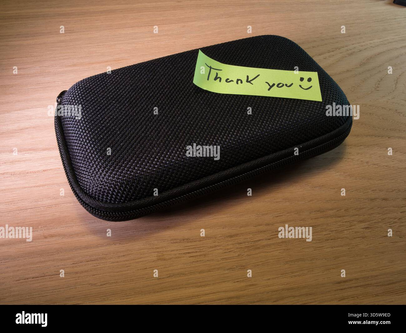 Nota di ringraziamento accanto alla custodia rigida nera - nota di ringraziamento scritta a mano con un viso sorridente accanto a una custodia rigida nera con cerniera su una scrivania di legno chiaro, co Foto Stock