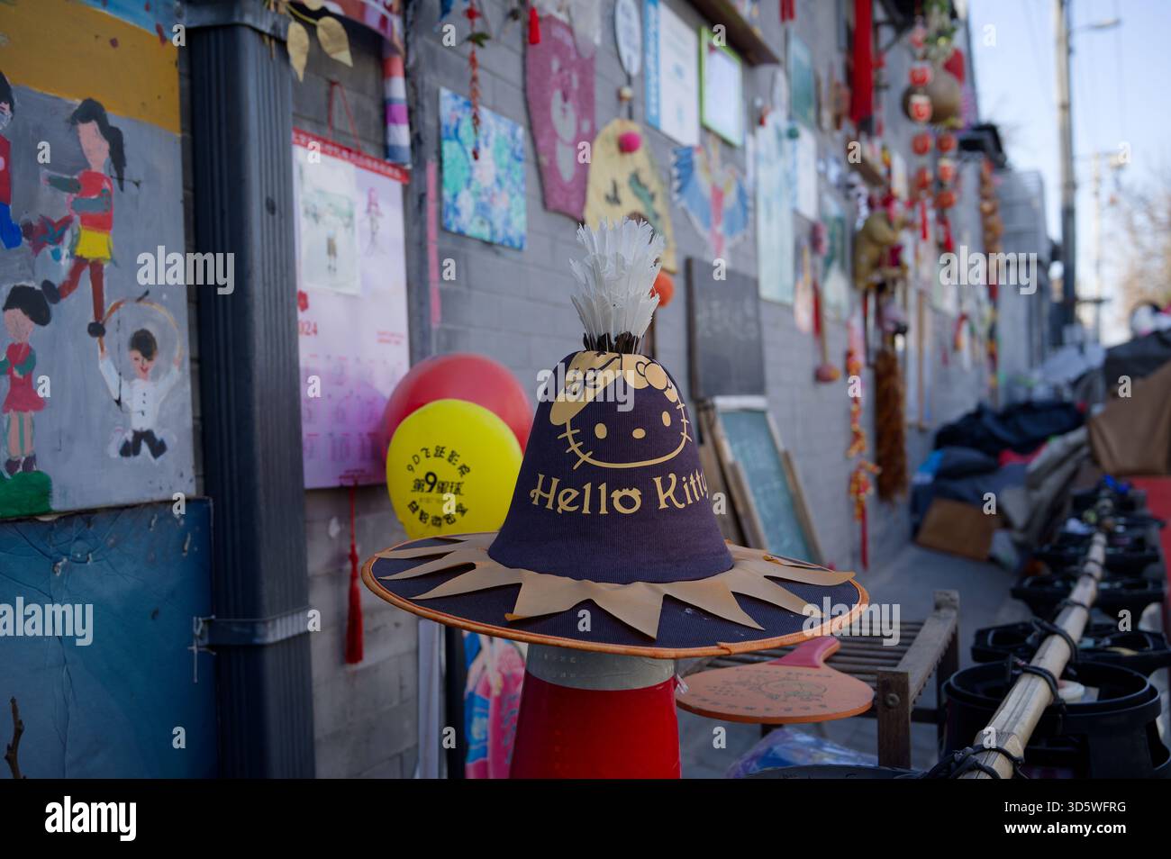 Cappello Hello Kitty appollaiato su un cono stradale fuori da una casa hutong, accanto a un muro di disegni per bambini a Pechino Foto Stock