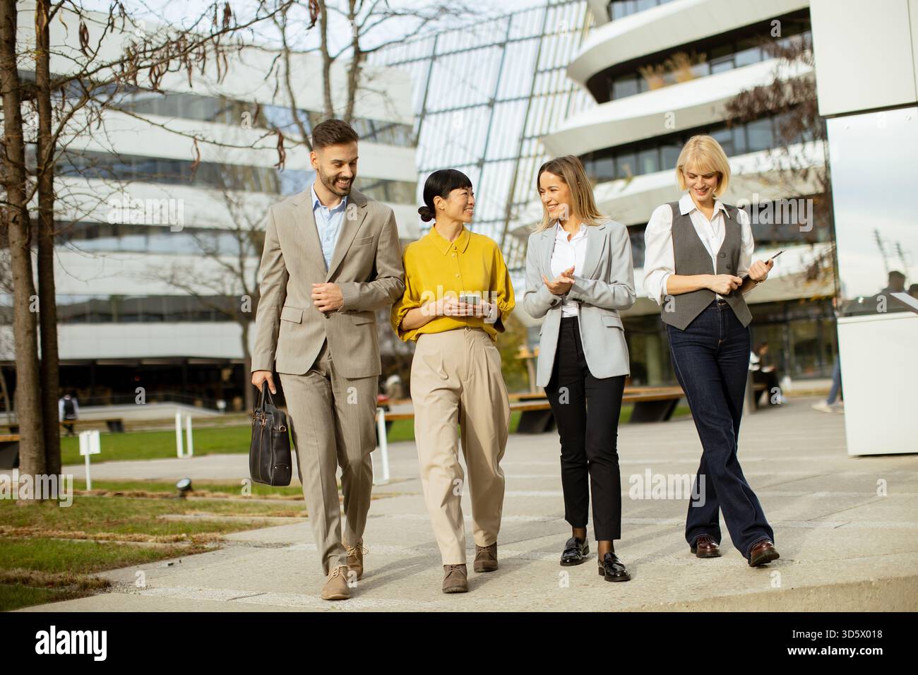 Quattro professionisti in abbigliamento da lavoro passeggiano insieme in un'area urbana. Sembrano coinvolgenti e felici, mostrando lavoro di squadra e collaborazione. La modalità Foto Stock