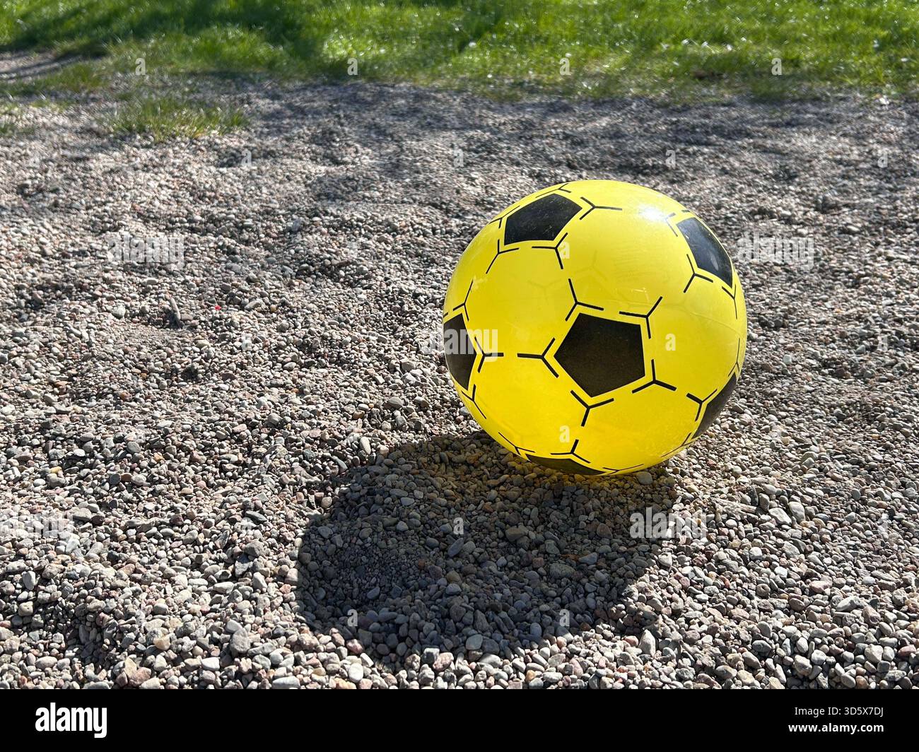 Un pallone da calcio giallo brillante con motivi neri appoggiato su una superficie ghiaia alla luce del sole - Immagine stock catturata con smartphone