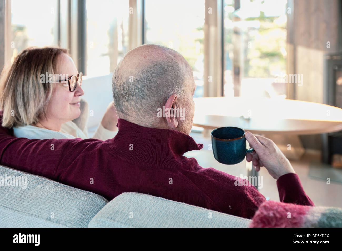 Una calda compagnia mentre una coppia senior si rilassa su un morbido divano nordico, condividendo una mattinata lenta con una conversazione tranquilla e una presenza gentile. Luce soffusa, c Foto Stock
