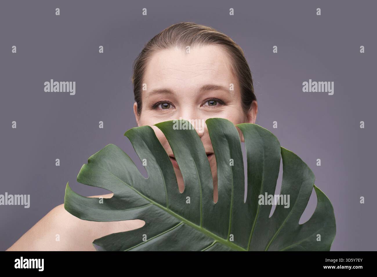 Donna adulta sorridente caucasica che sbircia dalla foglia di monstera su sfondo grigio Foto Stock