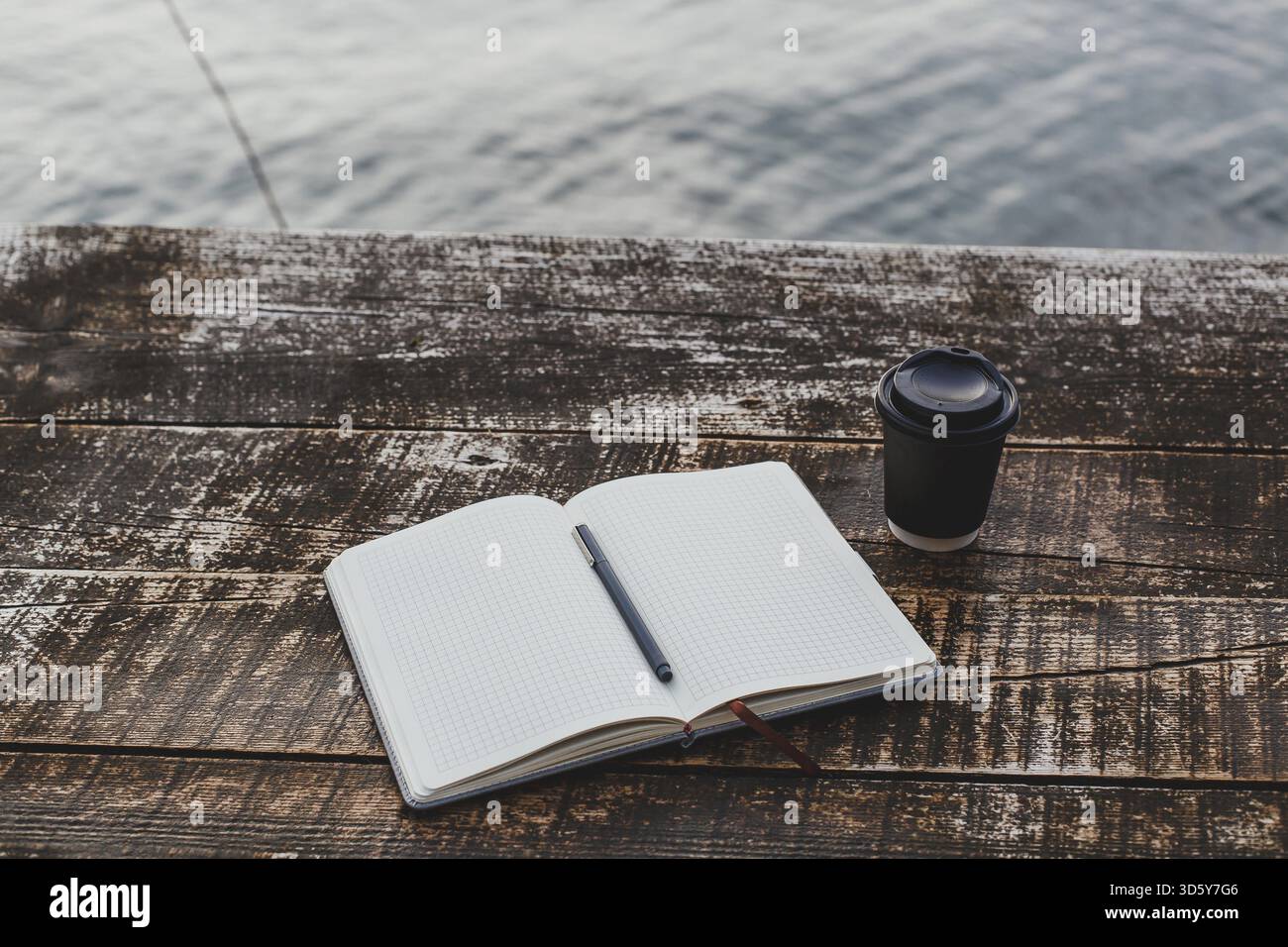 Un quaderno a griglia con una penna è aperto su un rustico tavolo in legno vicino all'acqua, accanto a una tazza da caffè da asporto nera, un ambiente tranquillo per scrivere o riscrivere Foto Stock