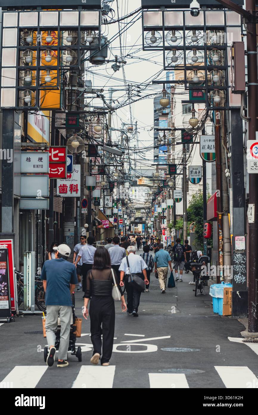 Le persone camminano lungo una strada trafficata e stretta fiancheggiata da grovigli di cavi, cartelli vintage e architettura unica in un vivace quartiere di Osaka. Foto Stock