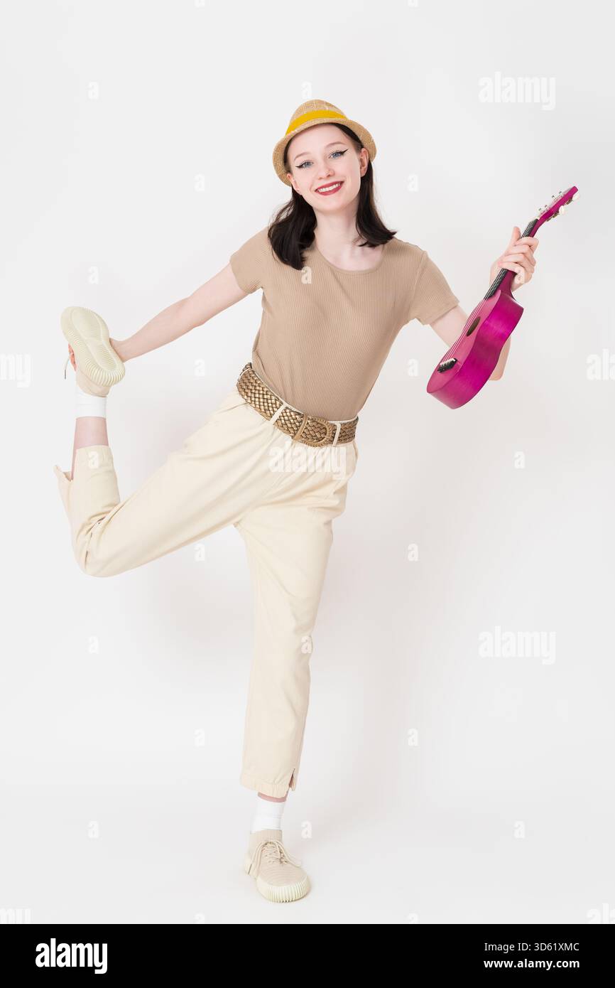 La giovane giocatrice di ukulele che emette un fascio di luce è in piedi su una gamba, ukulele in mano, caviglia tenuta con un sorriso giocoso. Una musicista donna positiva a tutta lunghezza sembra di Foto Stock