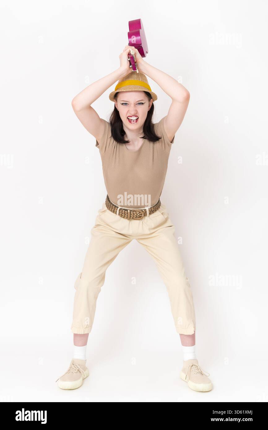 La musicista donna arrabbiata suonatrice di ukulele con un sinistro sorriso sulla faccia fa oscillare il suo ukulele sulla testa, pronto a colpire o distruggere lo strumento musicale in rabbia, fru Foto Stock