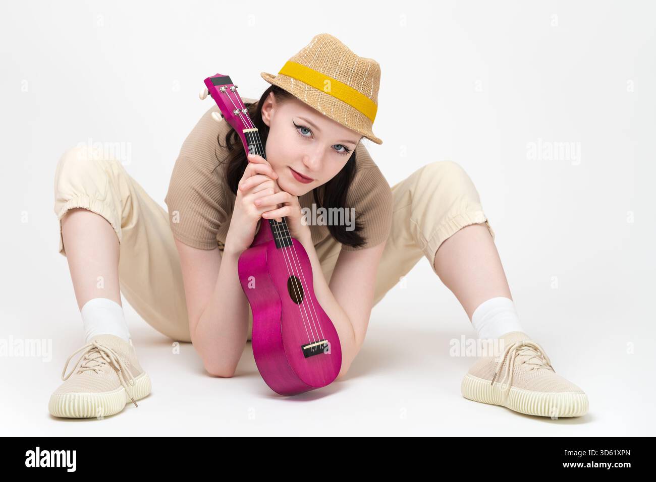 Una musicista con l'ukulele tra le mani si siede sul pavimento con le gambe separate e uno strumento musicale tra loro, appoggiandosi su di esso e guardando la telecamera W. Foto Stock