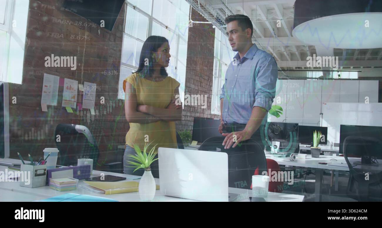 Discutere di colleghi che indossano camicia blu e camicetta di senape in un ufficio open space, con notebook bianco Foto Stock
