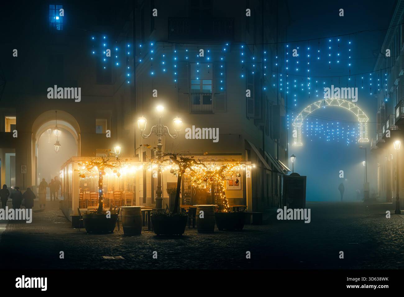 Le calde luci dorate di un ristorante all'aperto si illuminano di Natale in una notte invernale nebbiosa nel centro storico di Asti, in Italia. Foto Stock
