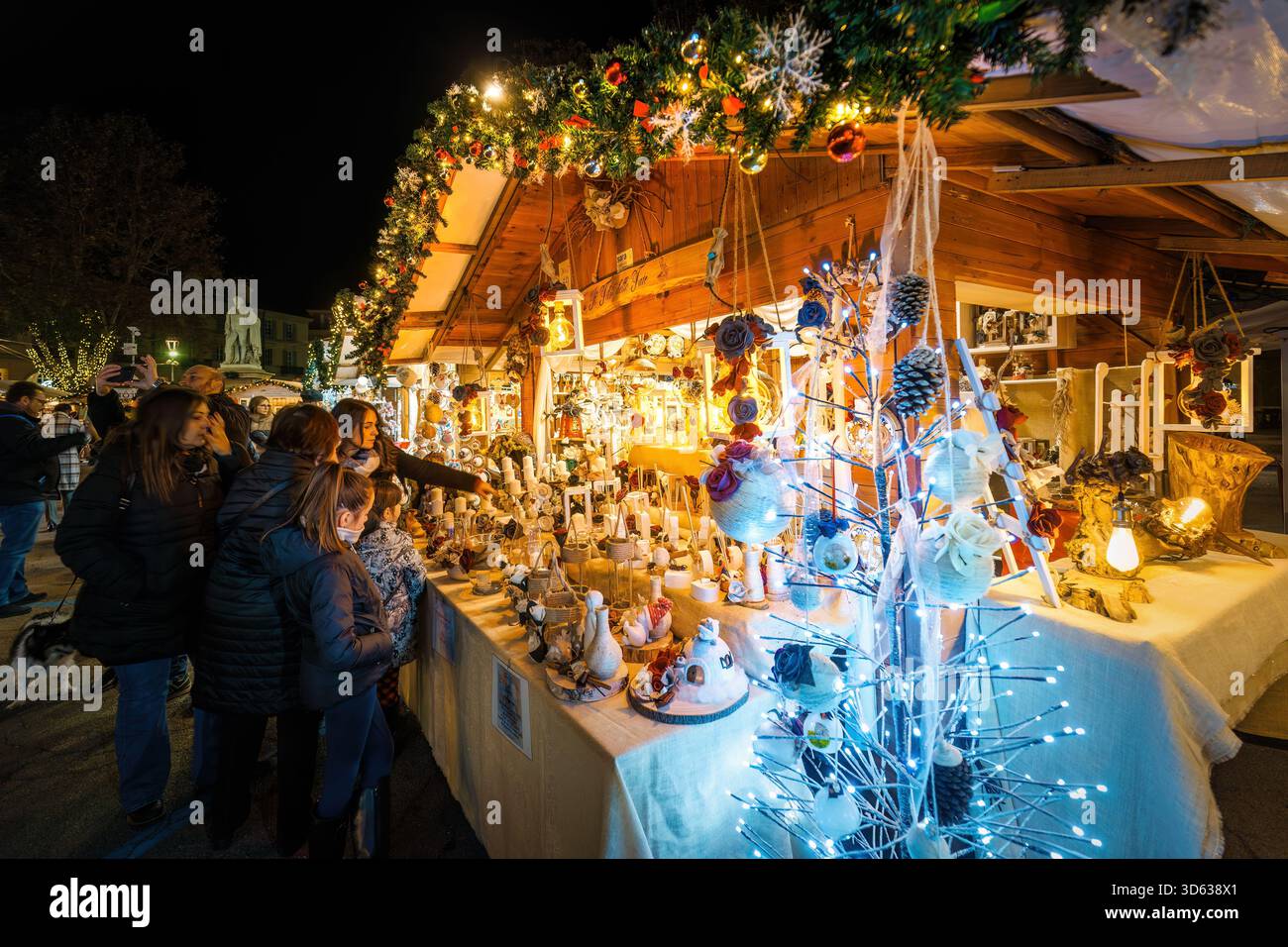 Un gruppo di acquirenti, illuminato da un caldo bagliore festivo, esamina gli ornamenti e le decorazioni fatti a mano esposti al mercatino di Natale di Asti. Foto Stock