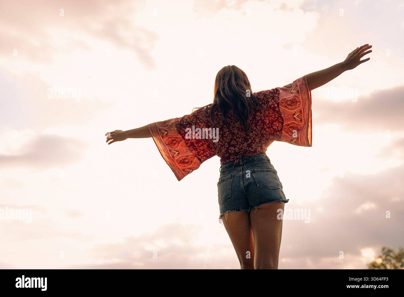 Donna con le braccia allungate che indossa abiti casual, rivolta verso l'esterno, che abbraccia l'aria fresca all'aperto sotto un cielo sereno. Concetto di libertà e stile di vita. Foto Stock