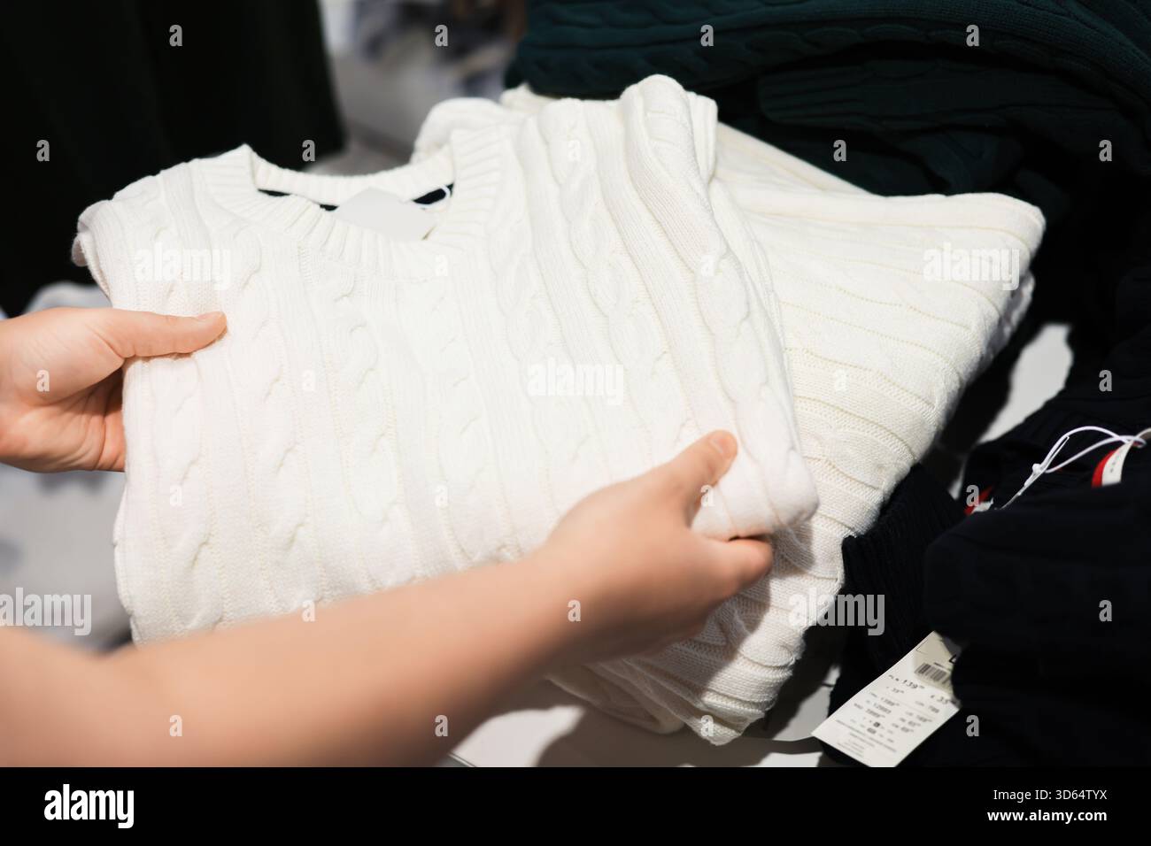 Le mani di una persona tengono in mano un maglione in maglia bianca piegato in un negozio di abbigliamento, con altri indumenti visibili nelle vicinanze. Foto di alta qualità Foto Stock