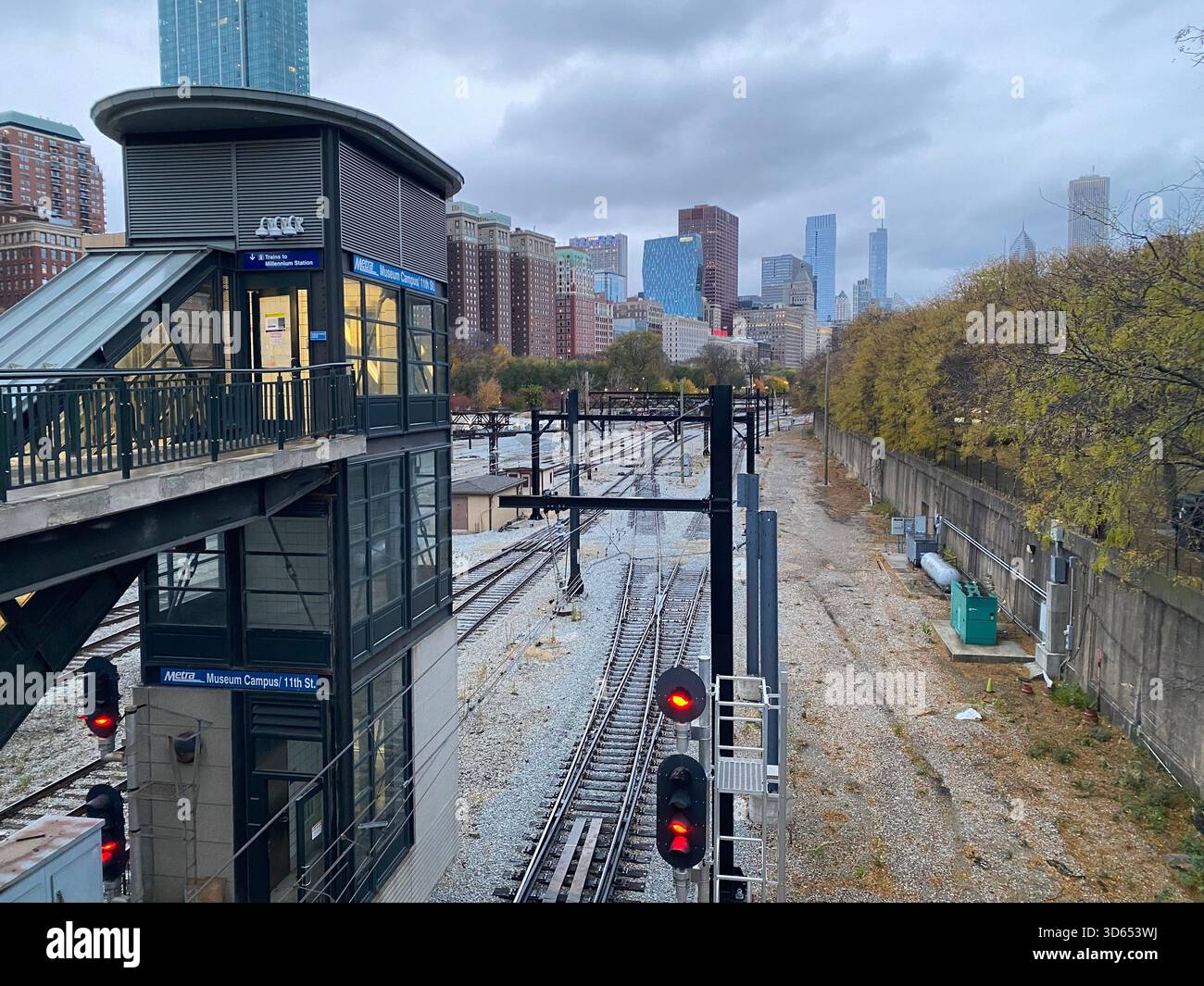 Una moderna stazione ferroviaria sopraelevata si affaccia su più binari ferroviari che conducono verso lo skyline del centro sotto un cielo nuvoloso. Gli alberi autunnali si allineano da un lato. - Immagine stock catturata con smartphone