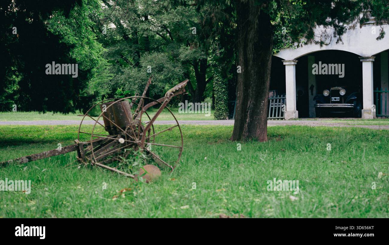 Vecchia attrezzatura agricola arrugginita con auto d'epoca e edificio rurale Foto Stock