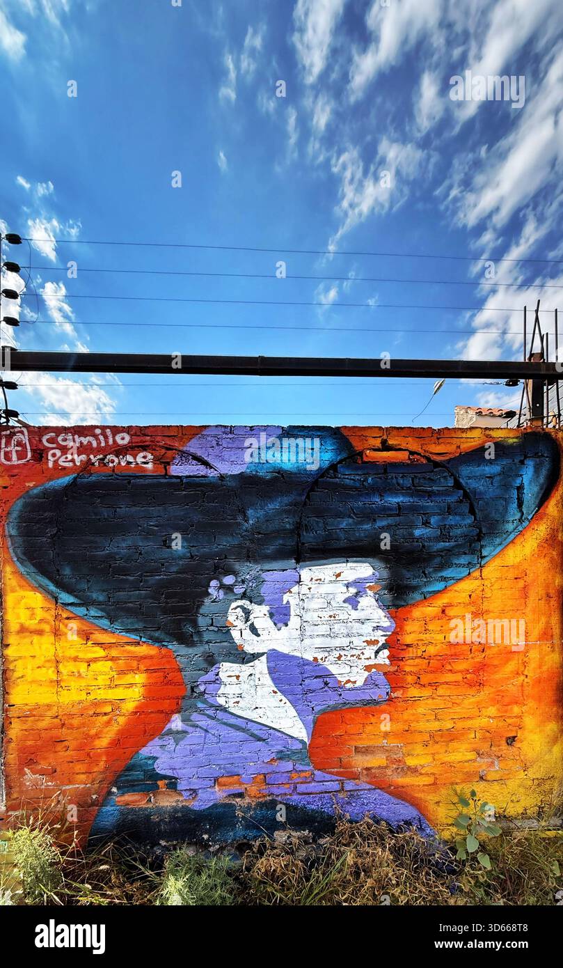 Colorato murale di un uomo in un cappello con un vivido sfondo arancione dipinto su una parete di mattoni sotto un cielo blu brillante con nuvole sparse - Immagine stock catturata con smartphone