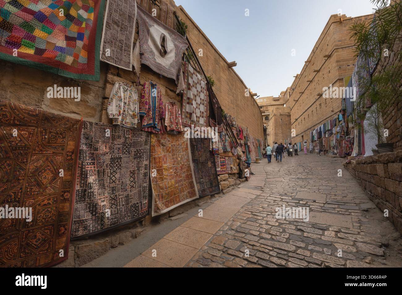 Tessuti colorati in un mercato di strada lungo uno stretto vicolo acciottolato all'interno del forte Jaisalmer, Rajasthan, con i visitatori che camminano attraverso. Foto Stock