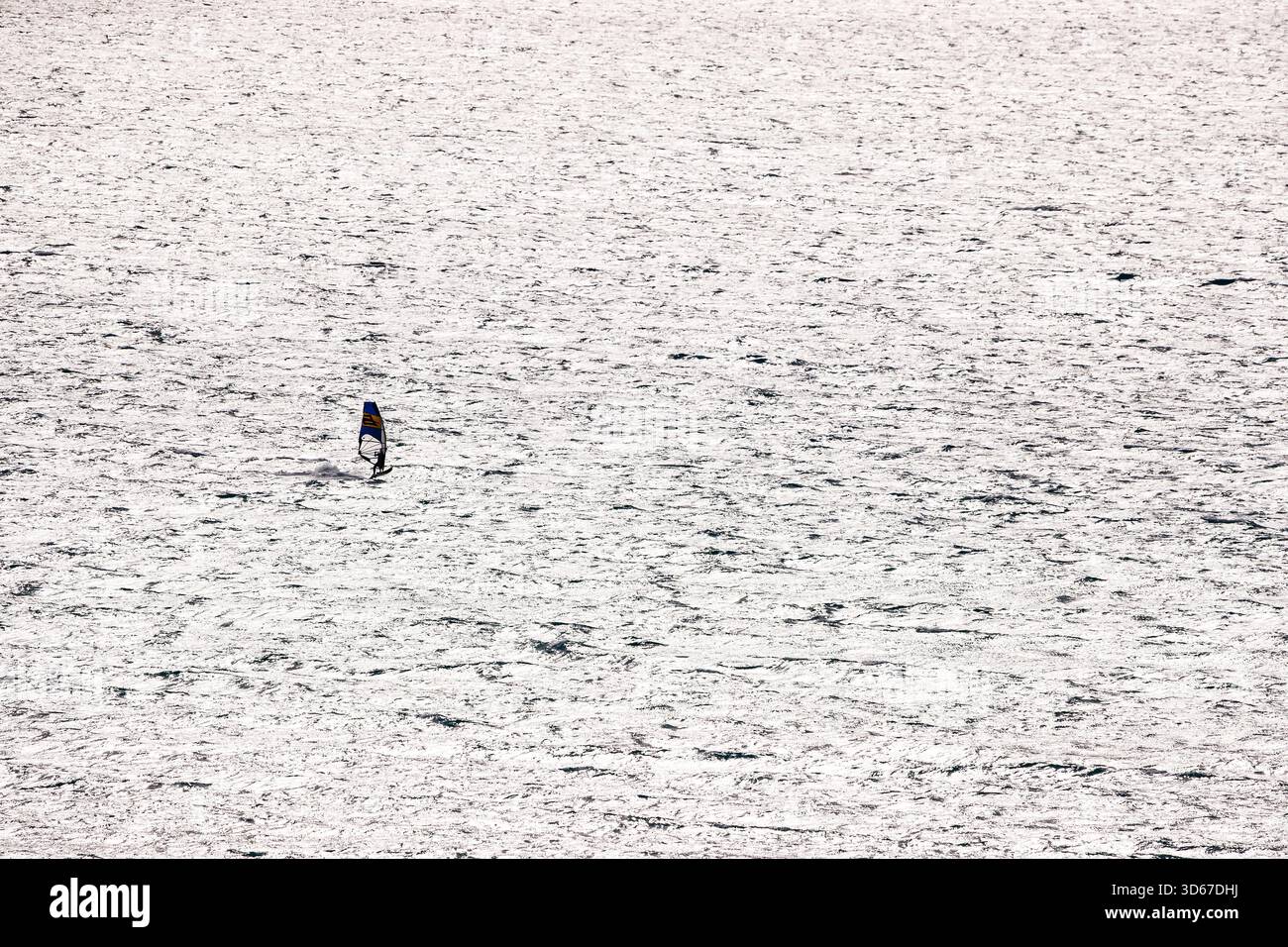 Un solo windsurfista con una vela blu e gialla è centrato nella vasta, intensa e strutturata distesa del Mar Mediterraneo che riflette il sole Foto Stock