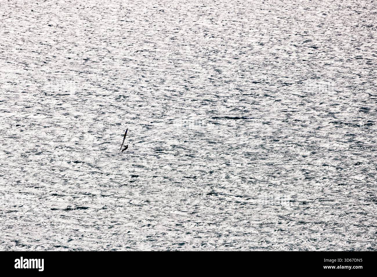 Una composizione minimalista che mostra un windsurfista solitario, quasi ridotto a una silhouette scura, che scorre orizzontalmente attraverso l'immensa luminosità e il testo Foto Stock