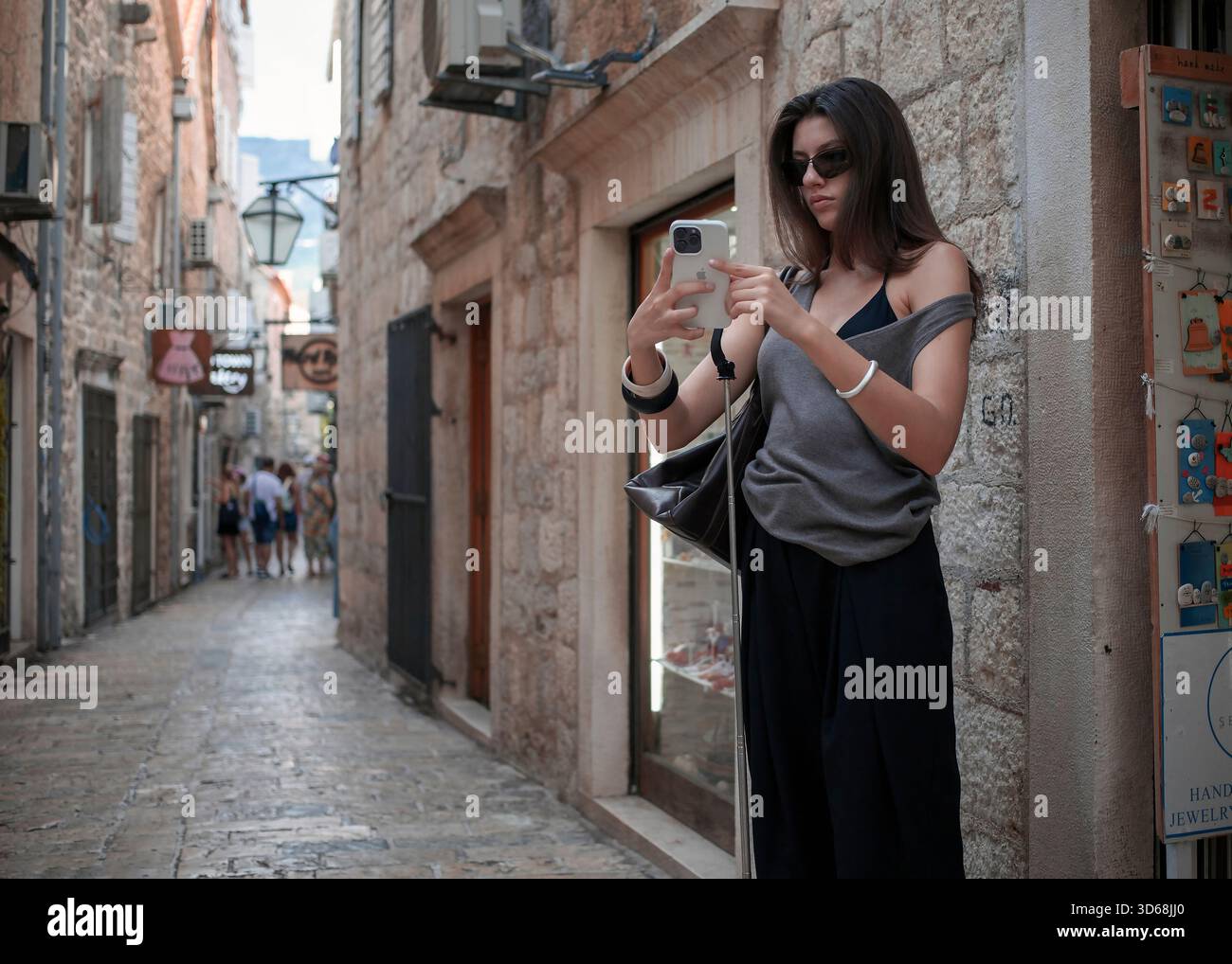 Giovane donna che usa lo smartphone in una stretta strada acciottolata nella città vecchia di Budva, in Montenegro, in un piacevole pomeriggio di fine estate. Foto Stock