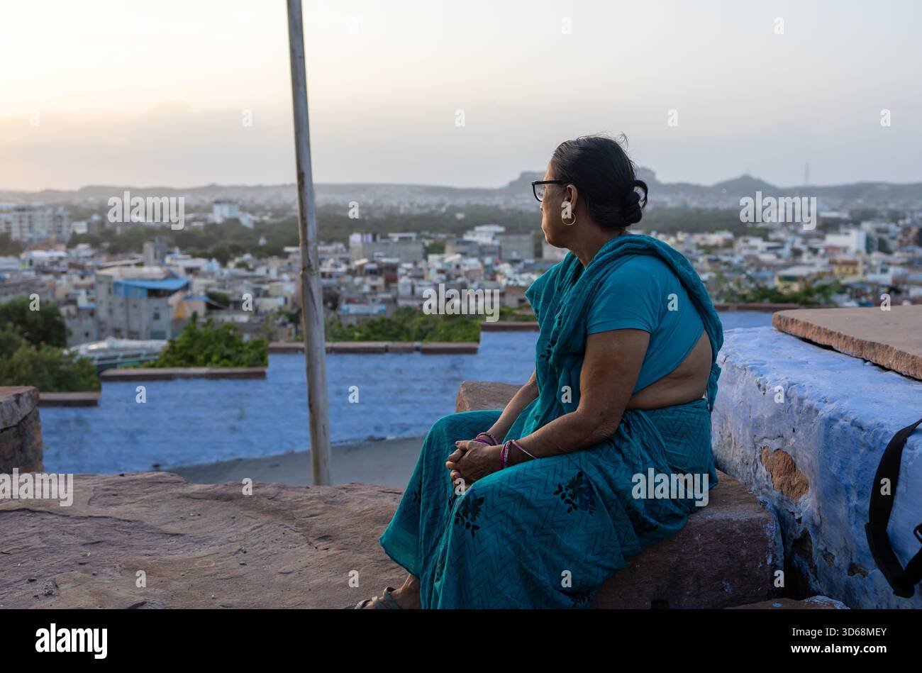 Anziana donna indiana in sari blu che guarda sullo skyline della città all'alba Foto Stock