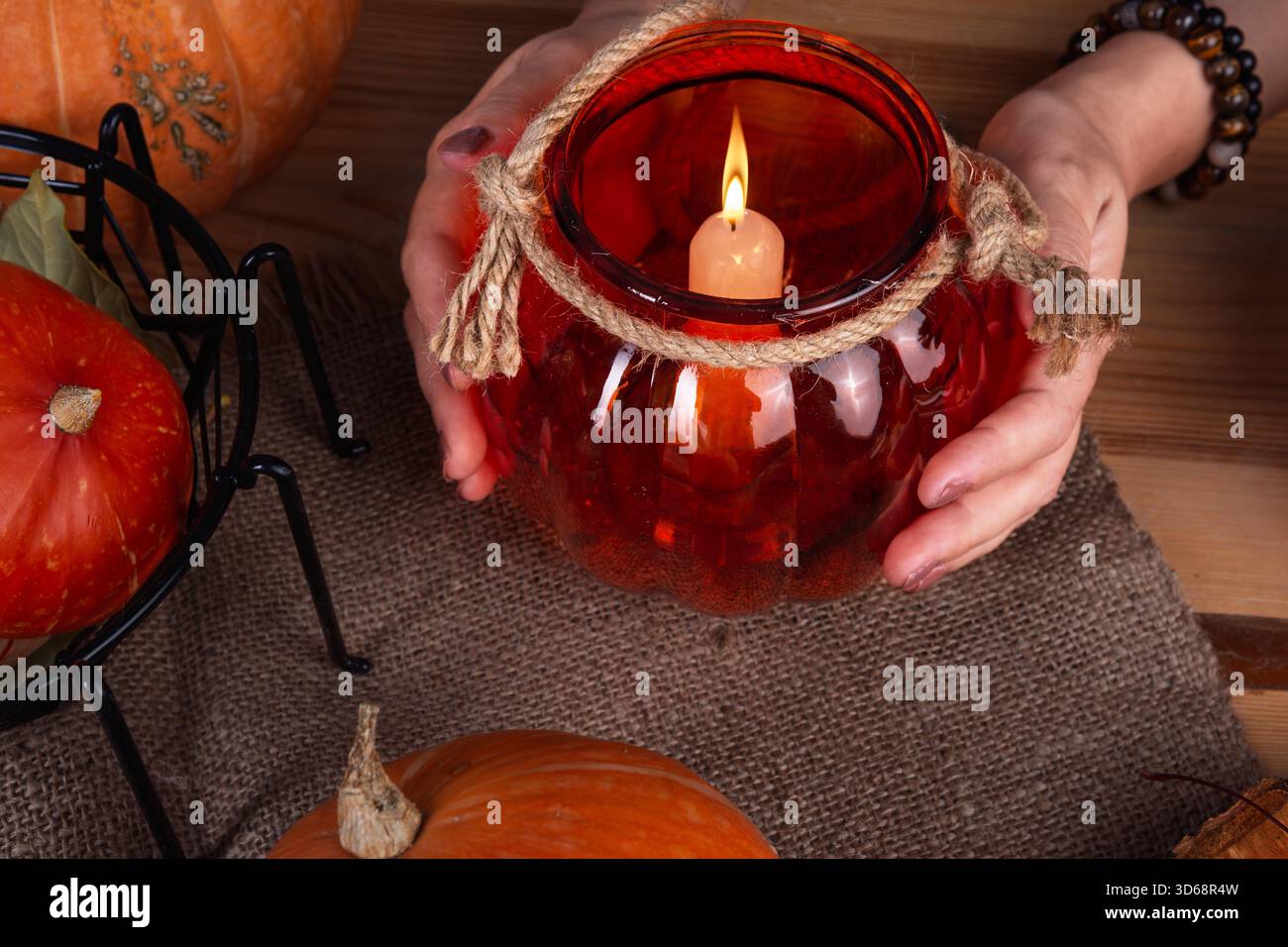 Una persona tiene in mano un portacandele decorativo a forma di zucca con una candela accesa all'interno. Una zucca arancione è collocata nelle vicinanze su un tavolo di legno Foto Stock