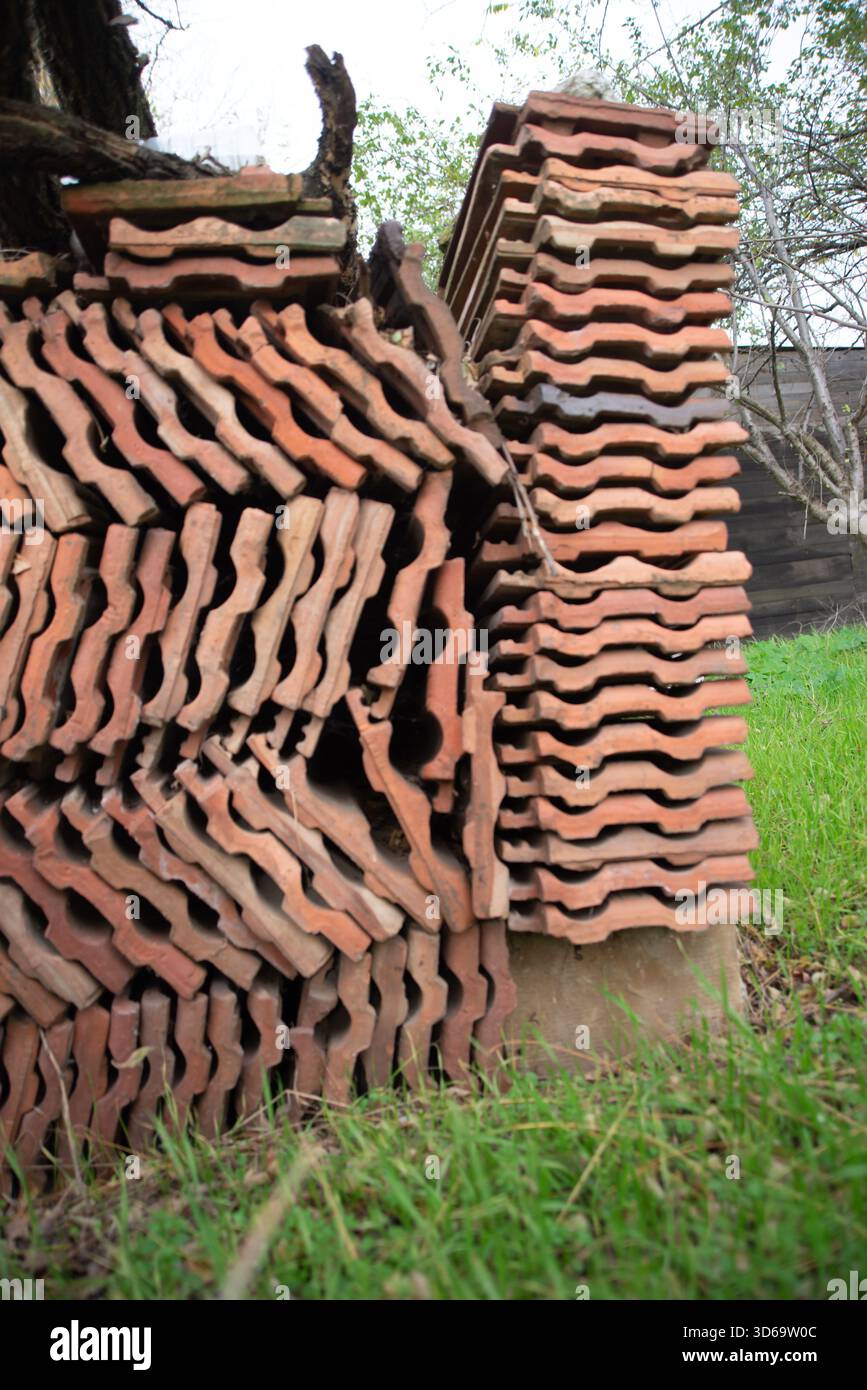 La foto mostra un'alta pila irregolare di tegole di terracotta disposte a strati densi accanto a un albero, con erba verde in primo piano e rami Foto Stock