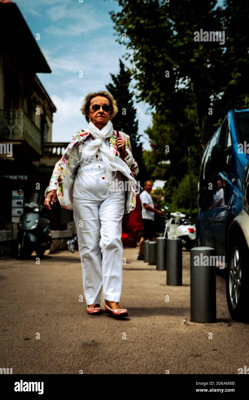 Donna elegante che cammina lungo una strada soleggiata a Nizza, in Francia, catturata in un momento schietto con alberi, auto e dettagli urbani sullo sfondo. Foto Stock