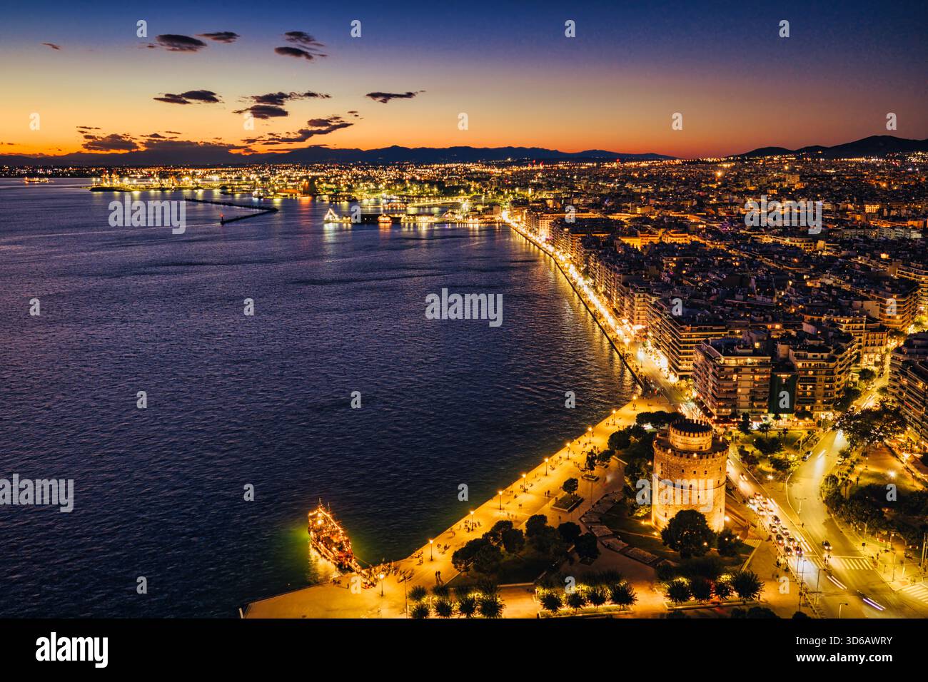 La Torre Bianca si illumina mentre il crepuscolo abbraccia Salonicco. Le luci risplendono sul mare, un molo festoso e la città respira storia, movimento e M Foto Stock