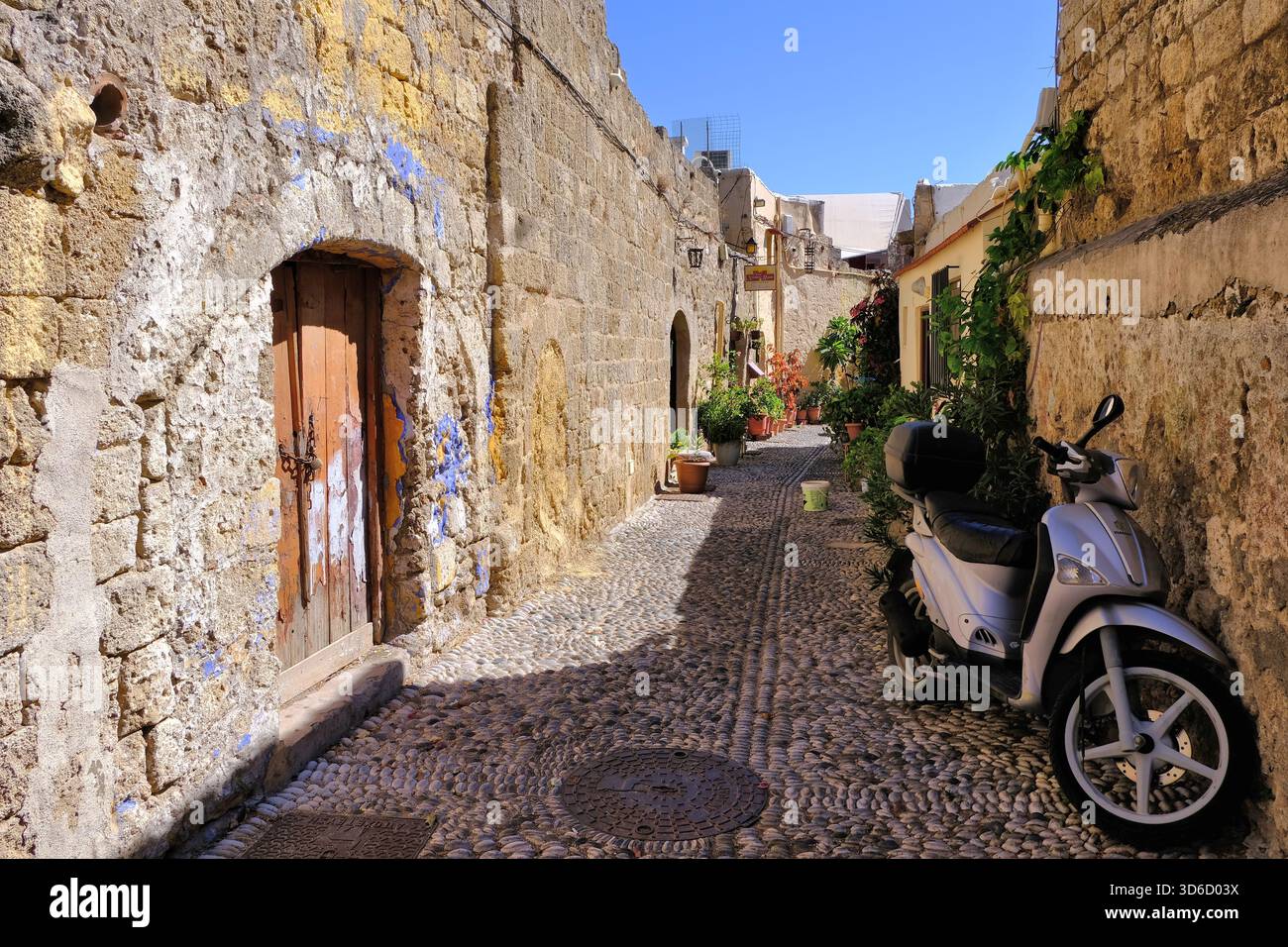 Città vecchia di Rodi: Strada curva di Klisthenous con porte ad arco, sentiero di ciottoli, tombino con cervi, fiori e moto a Rodi, Grecia Foto Stock