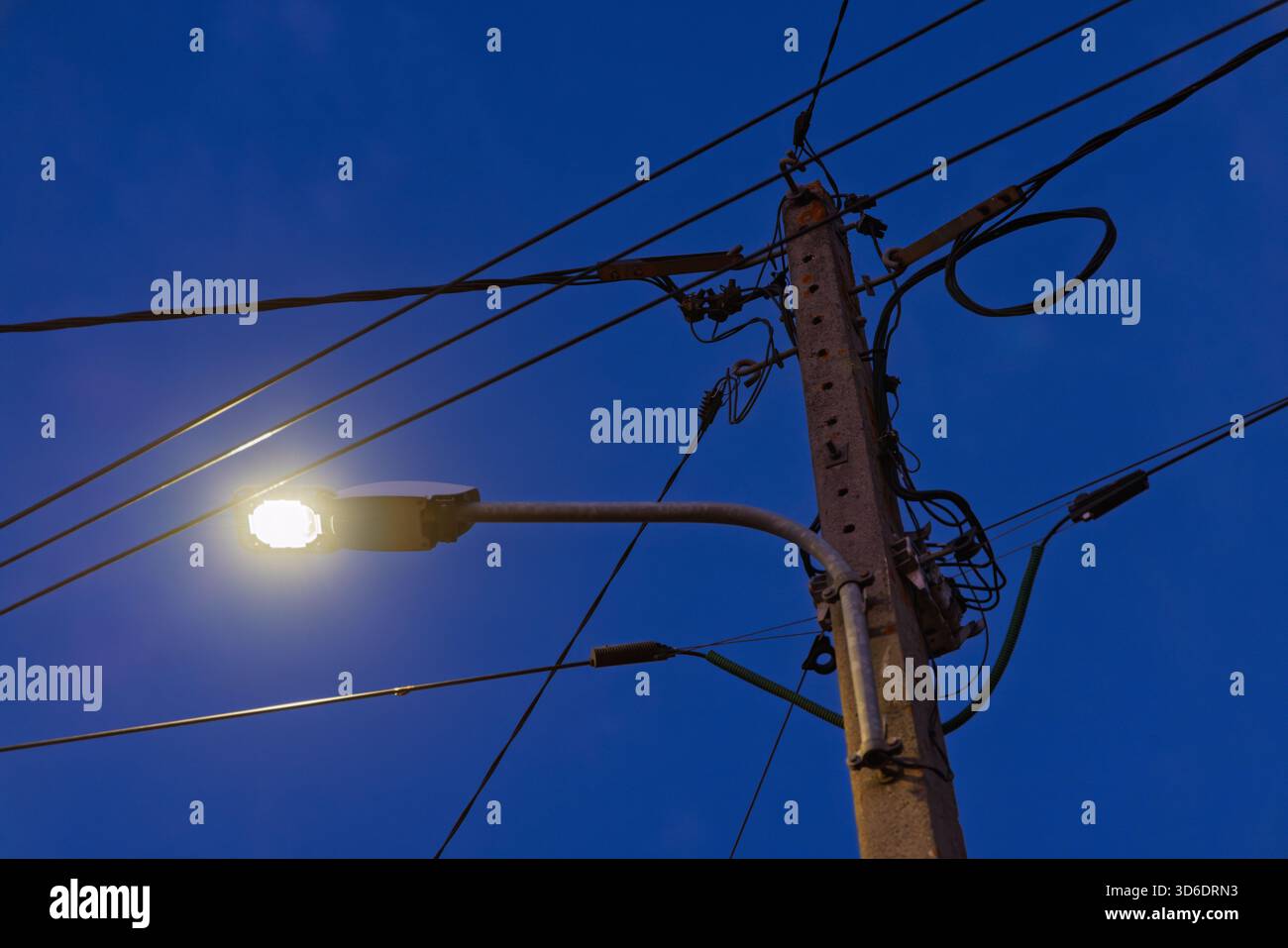 Il moderno lampione a LED brilla di luce al crepuscolo. Palo di servizio con fili complessi contro un cielo notturno blu profondo. Infrastruttura ed energia. Foto Stock