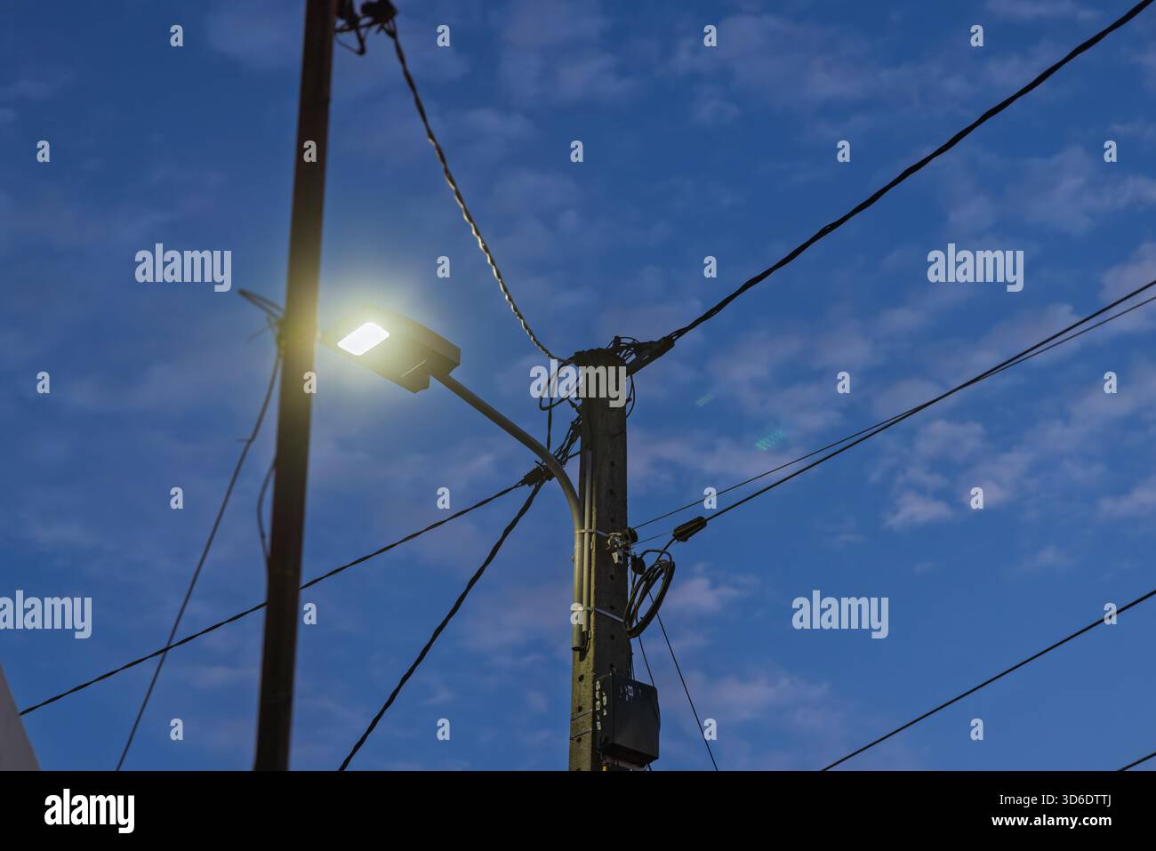 Una luce LED illuminata collegata a un palo in cemento, che splende in modo luminoso contro un cielo notturno blu intenso con nuvole morbide e molti fili. Foto Stock