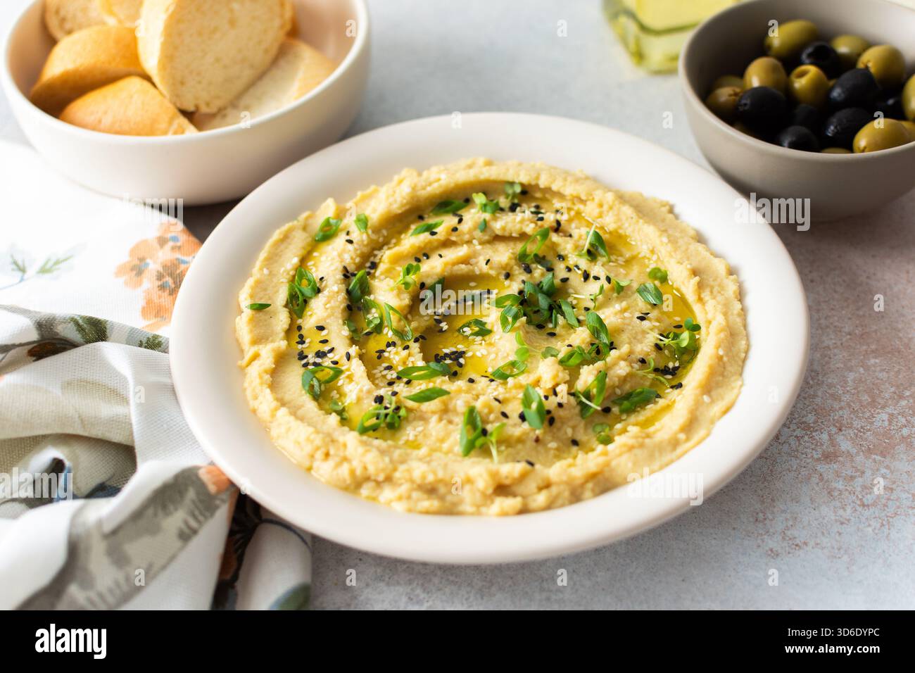Una ciotola di hummus cremoso ricoperta di olio d'oliva e erbe aromatiche si trova accanto al pane fresco e a una ciotola di olive. L'illuminazione è caratterizzata da un design semplice e invitante Foto Stock