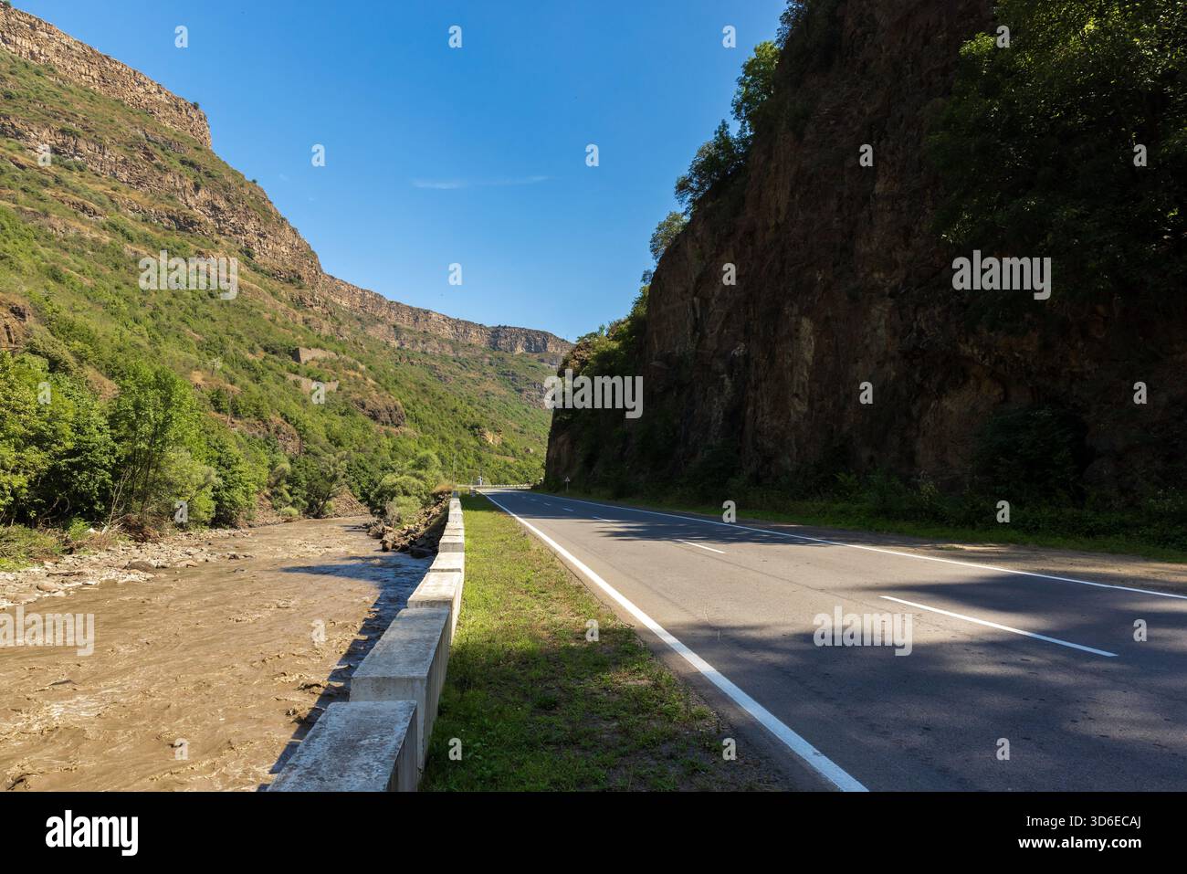 Una strada tortuosa attraversa un canyon, fiancheggiata da alte pareti rocciose e vegetazione lussureggiante. Un fiume poco profondo scorre accanto ad una barriera sotto un cielo azzurro, evoki Foto Stock