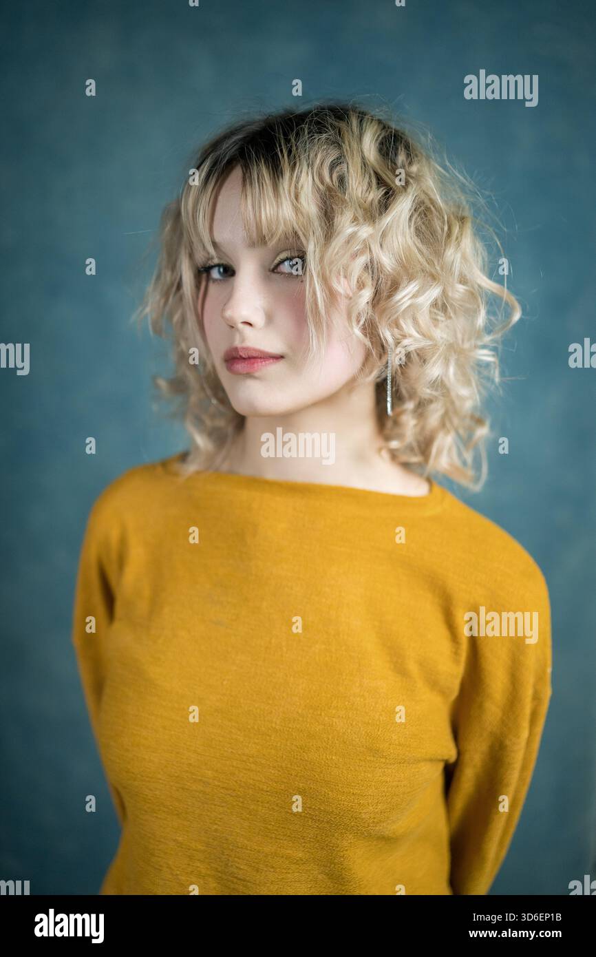 Una giovane donna con capelli biondi ricci si posa su uno sfondo blu, indossando un top giallo senape. La sua espressione sicura e il suo abbigliamento casual creano un Foto Stock