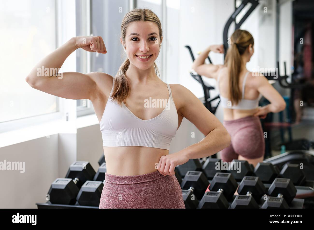 Una donna sorridente si flette il bicipite, ammirando il tono muscolare nello specchio della palestra, irradiando salute e sicurezza Foto Stock