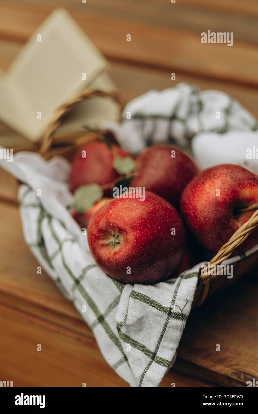 Mele rosse fresche e succose disposte in un cesto rustico intrecciato su un tavolo di legno, parzialmente ricoperte da un panno a scacchi verde e bianco. Foto Stock