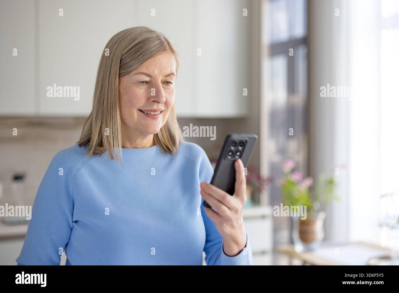 Donna anziana sorridente mentre tiene in mano e utilizza uno smartphone per una videochiamata, comunica con la famiglia o gli amici e resta connessa tramite la tecnologia Foto Stock