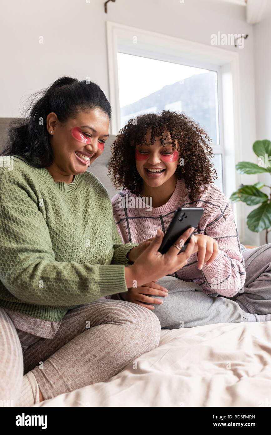 Madre e figlia condividono lo smartphone mentre indossano un divano e delle toppe in gel sul letto in camera da letto Foto Stock