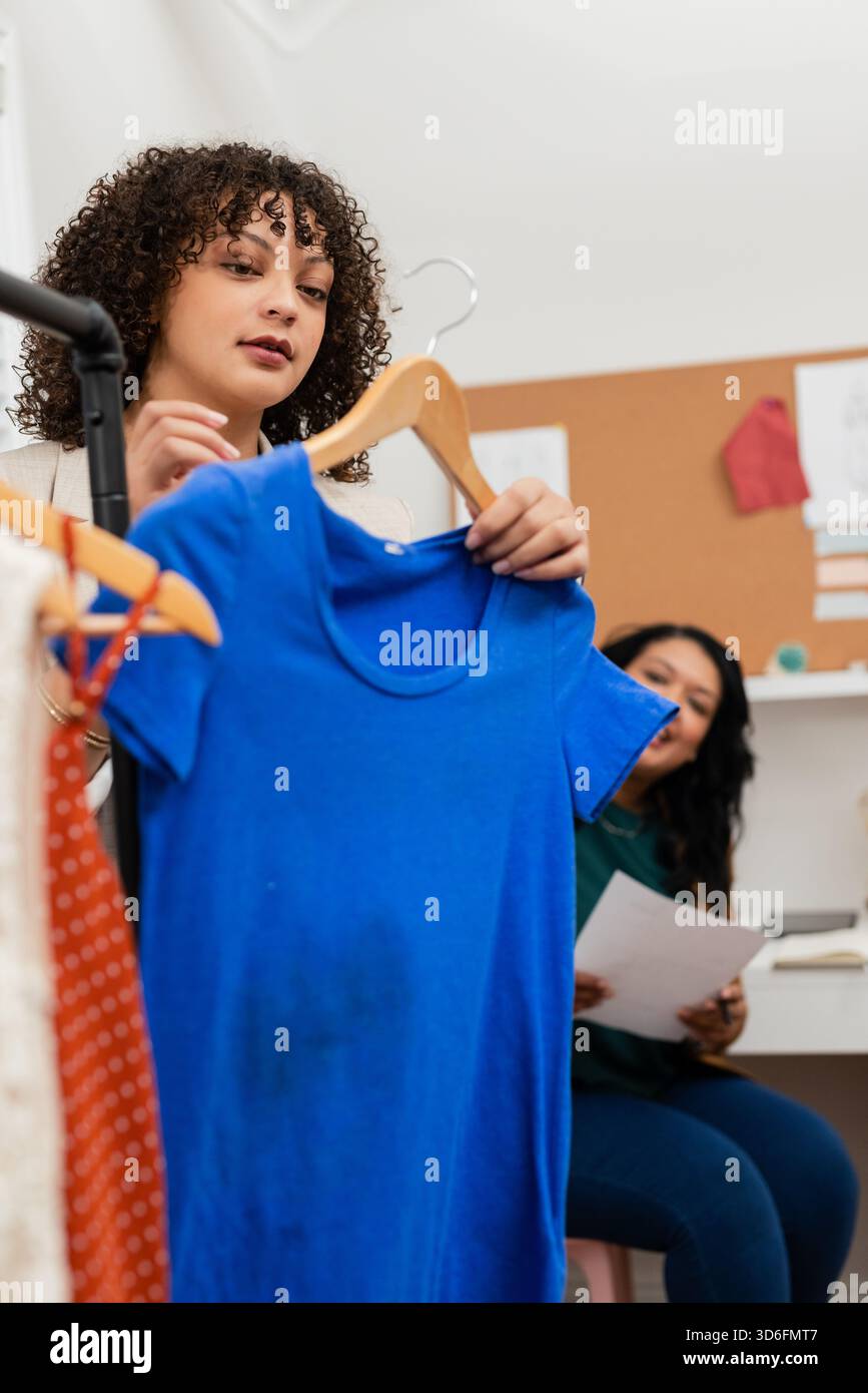 Diverse collaboratrici che tengono una t-shirt blu brillante su un appendiabiti, sedute alla scrivania nello studio di design Foto Stock