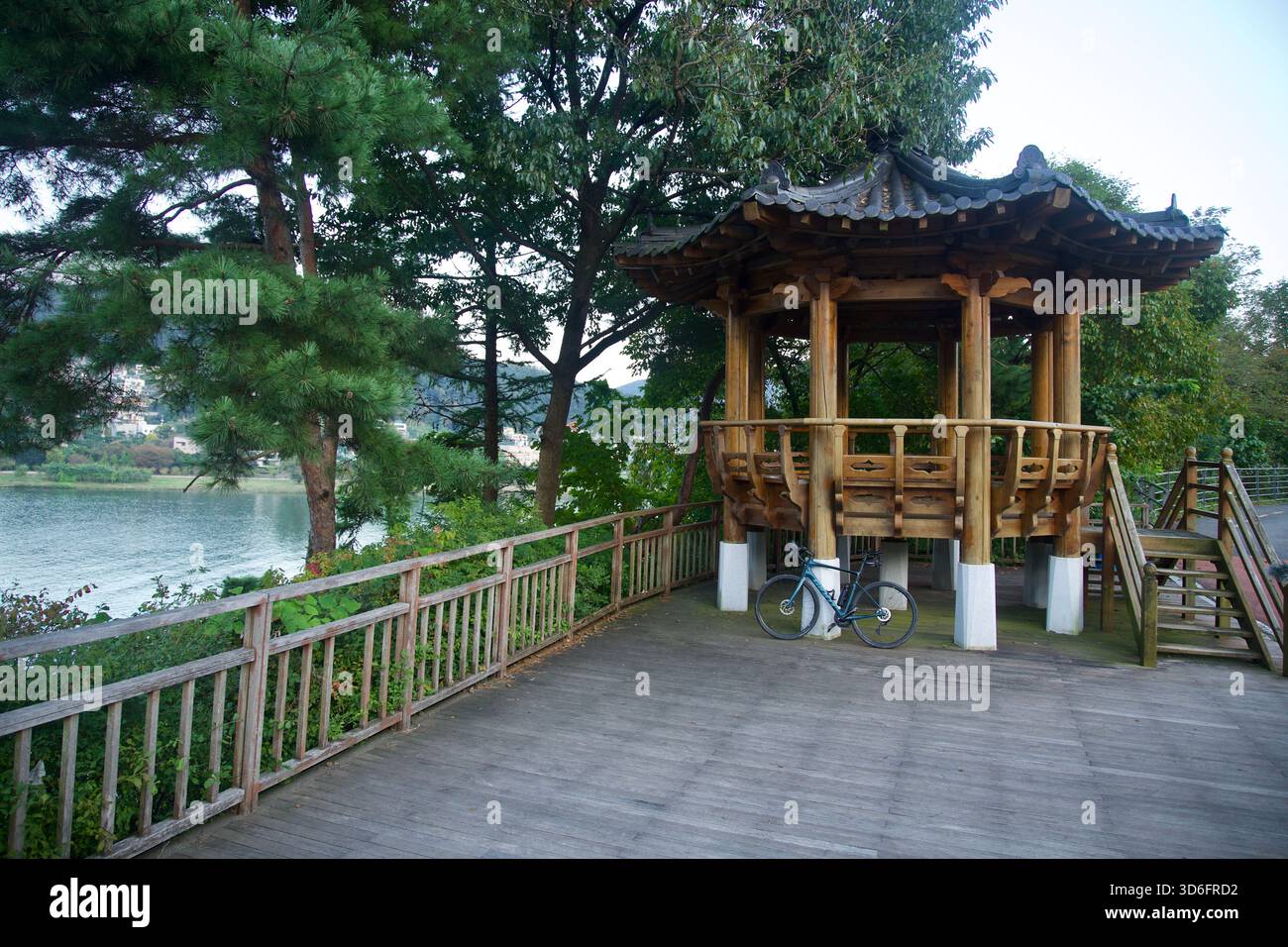 Un padiglione in legno lungo il fiume sorge accanto agli alberi di pino sul ponte di Bukhangang a Namyangju, con una bicicletta appoggiata su un palo e case sull'acqua Foto Stock