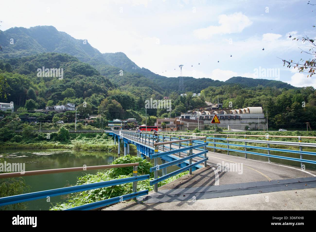 Un ponte blu porta la pista ciclabile attraverso una stretta insenatura del lago Uiam verso un piccolo villaggio, con cabine in collina e la linea della funivia visibile aga Foto Stock